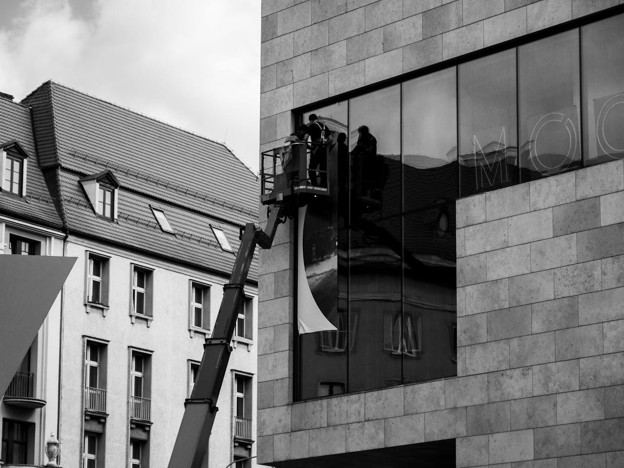 services-02 Black and white photo of workers using a lift to install a glass panel on a modern building exterior.